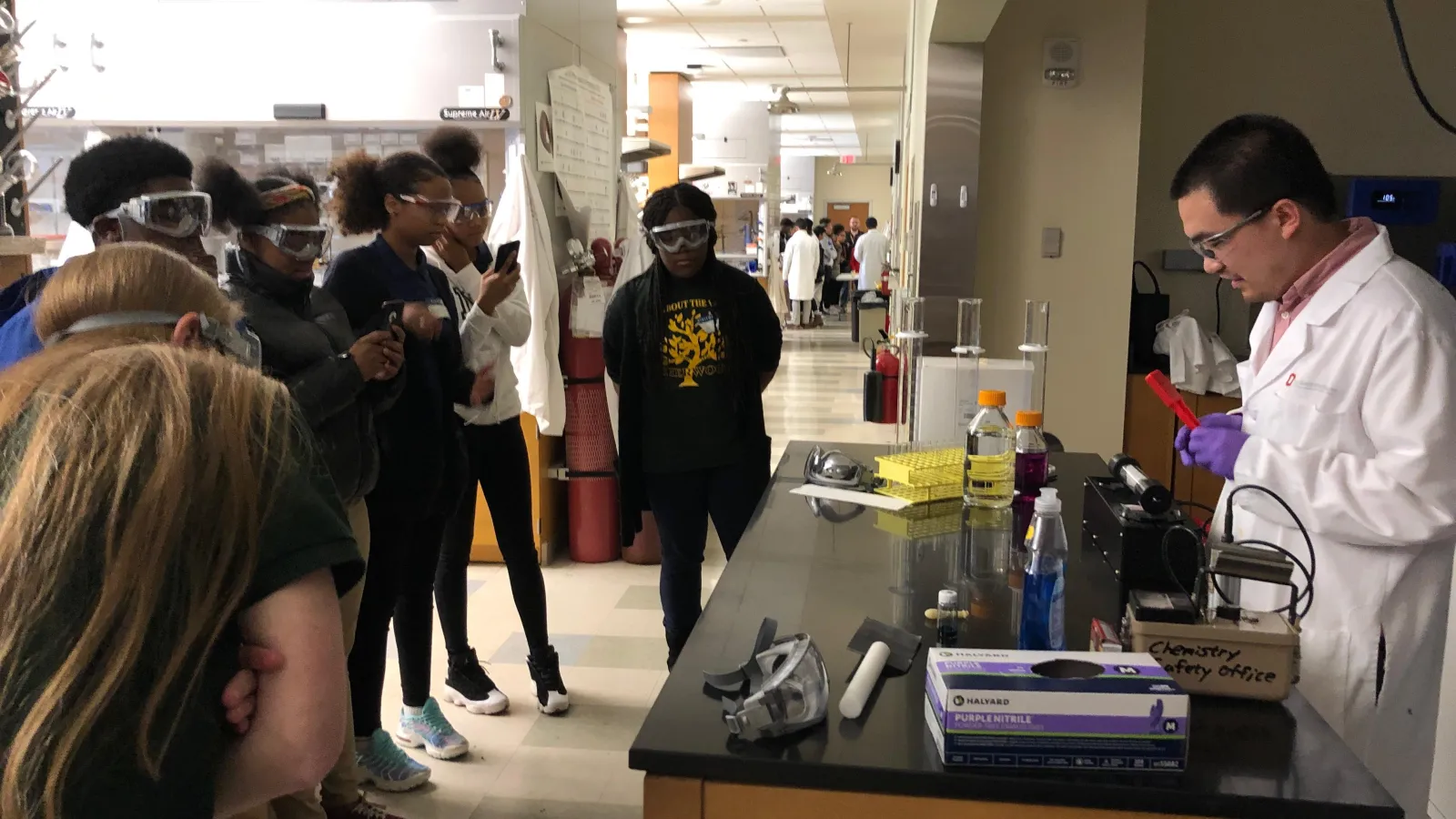Multiple students stand around a table wearing goggles while a chemistry demonstration is performed. 