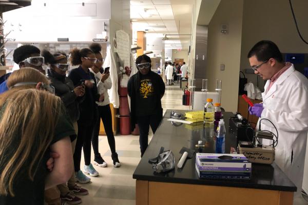 Multiple students stand around a table wearing goggles while a chemistry demonstration is performed. 